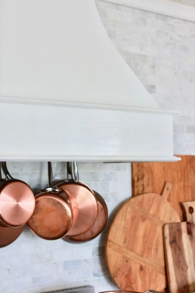 Copper pans hanging inside a white kitchen hood; wooden cutting boards are resting against the wall.