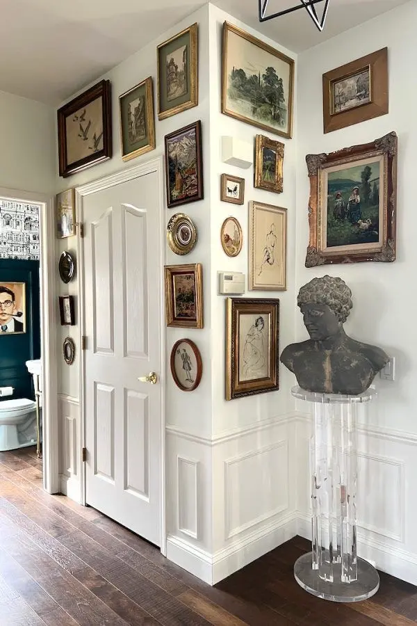 Classical paintings and drawings in various gilded frames hanging on a white wall with wainscoting; there’s a white door and a dark sculptural bust on a transparent pedestal.