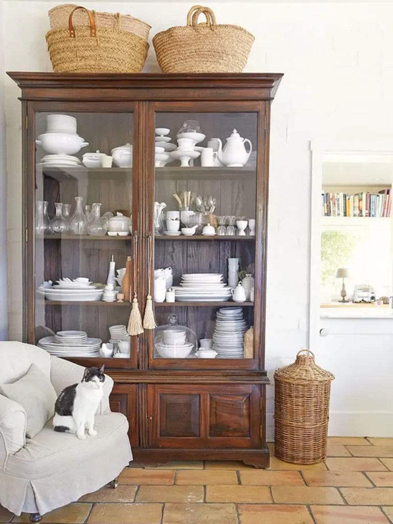 A large vintage wooden hutch displaying a collection of white tableware behind glass doors and three wicker baskets on top, a stone floor, a vintage wicker container, and a cat sitting on a gray armchair.