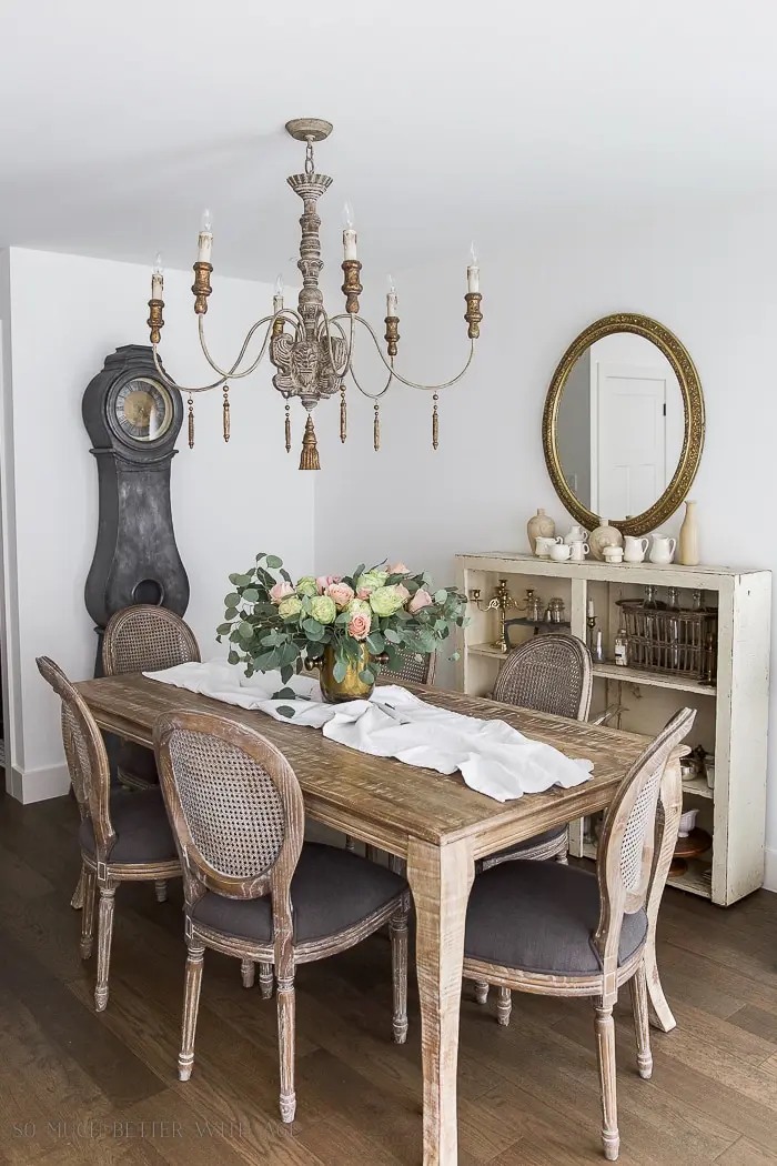 A vintage French dining room with a distressed grandfather clock, a rustic wooden table with cane-back chairs, a vintage French chandelier, a round gilded mirror, and a bouquet of fresh roses in a vase.