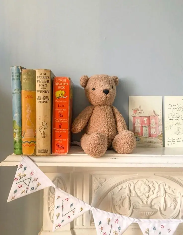 An ornate off-white mantel with four colorful children’s books, a teddy bear, postcard, and a vintage-style felt bunting.