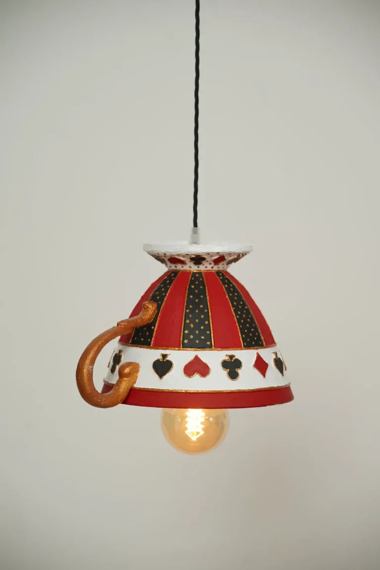 A hanging lamp made from a sculpture of an upside down teacup inspired by Alice’s Adventures In Wonderland, featuring a black, red, and white pattern with playing cards motifs.