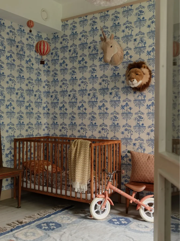 A vintage-inspired nursery with a wooden crib, a small kids’ bicycle, hot air balloon hanging decorations, plush unicorn and lion wall hangings, a vintage rug, and a British Colonial-inspired blue and white patterned wallpaper with a palm tree motif.