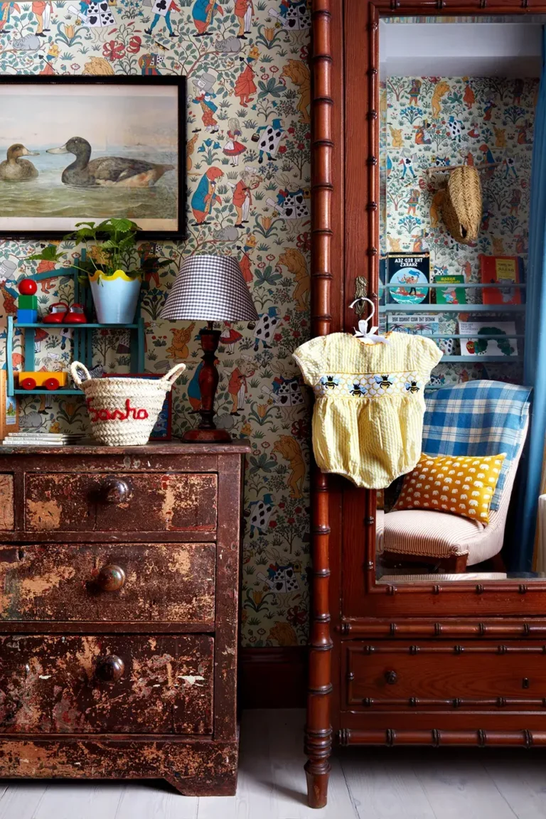 A vintage-inspired nursery with an old distressed dark wood dresser, an antique wooden hutch, a vintage-style Alice In Wonderland wallpaper, a gingham-patterned table lamp, an old-fashioned art print of mallards, and other baby toys and accessories.