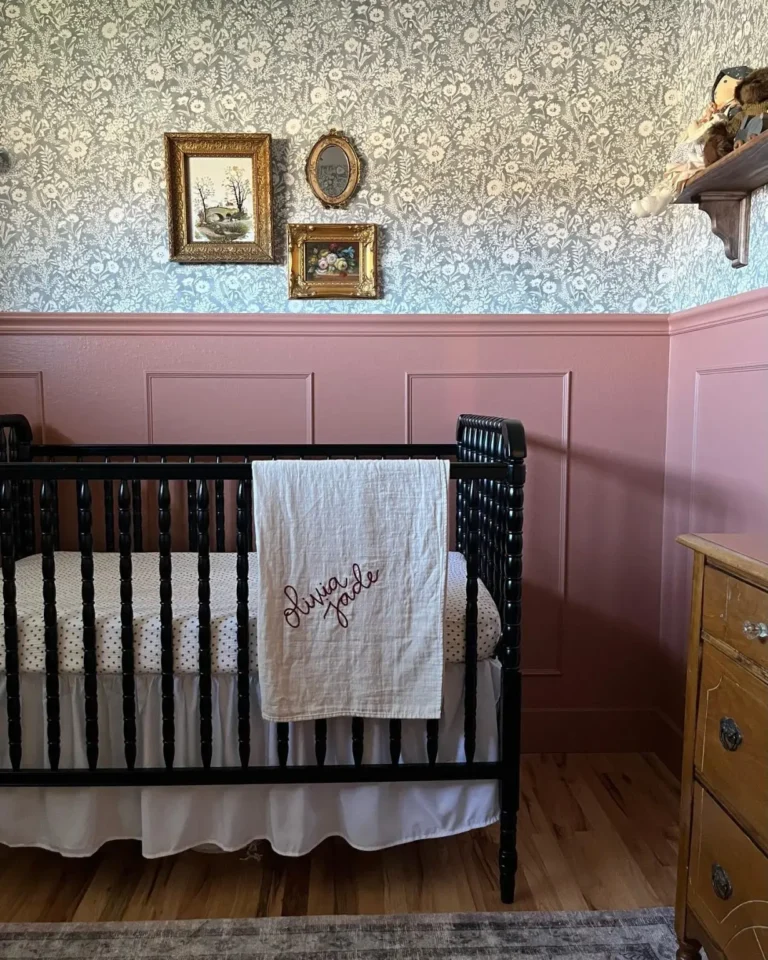 A vintage black crib in a vintage-inspired nursery with blush pink wainscoting and a William Morris wallpaper with three vintage-style gilded orange frames.