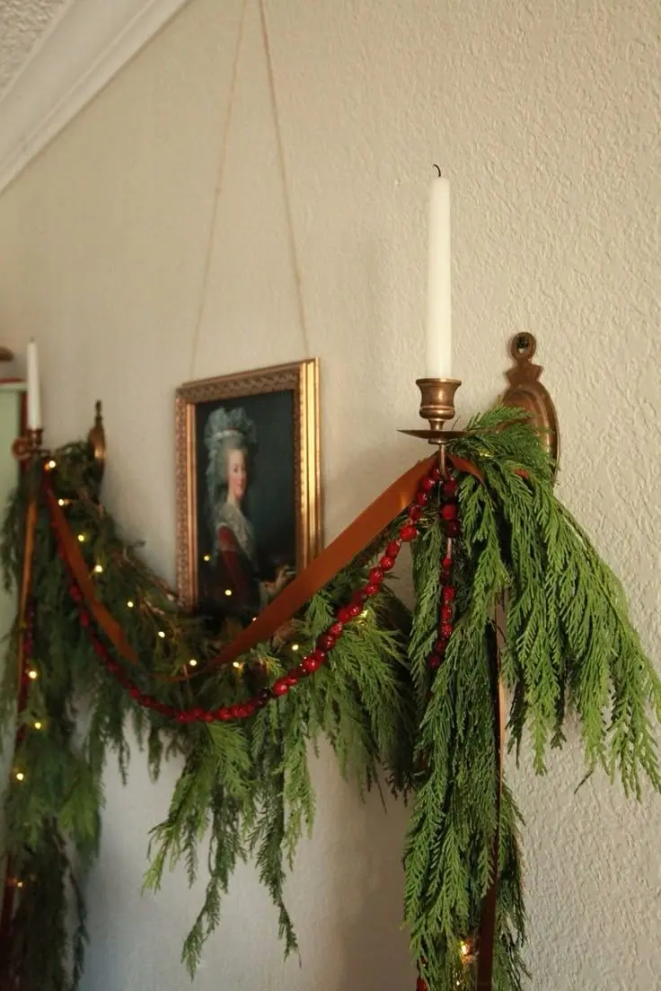 A garland made up of fresh fir, red beads, a brown ribbon, and tiny string lights draped over antique brass wall sconces with candlesticks. A 17th century painting in an ornate gilded frame hangs between the two sconces.