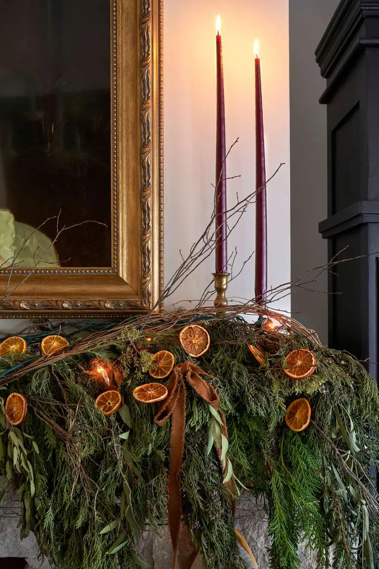 A Victorian-style Christmas garland made up of real holly, mistletoe, and fir, slices of orange, and rustic brown ribbons. Behind the garland is a mirror in an ornate gilded frame and two burgundy candlesticks in vintage brass candlestick holders.