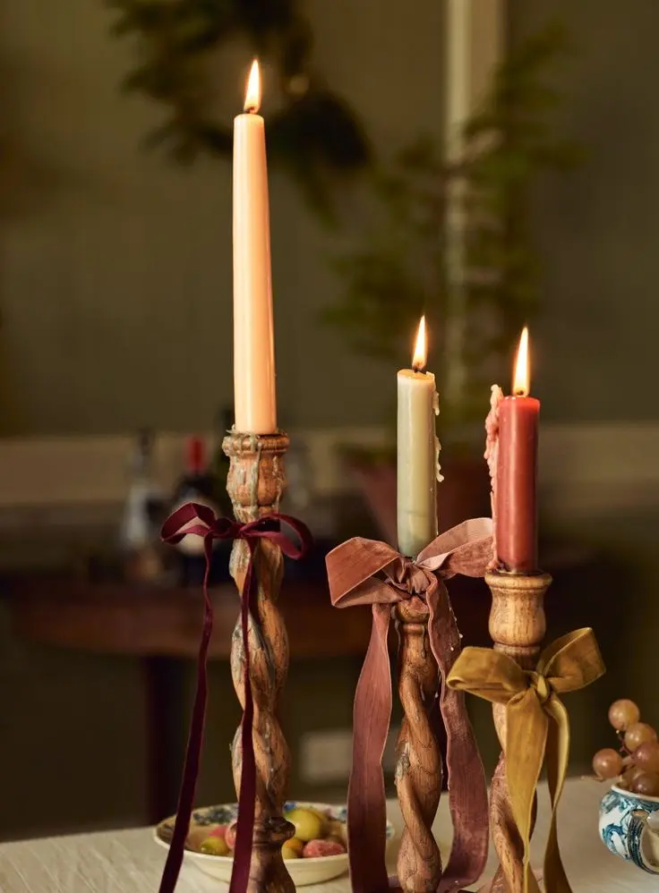 Three wooden candlestick holders with beige and rust-colored candlesticks sitting on a table with a fruit bowl seen in the background.