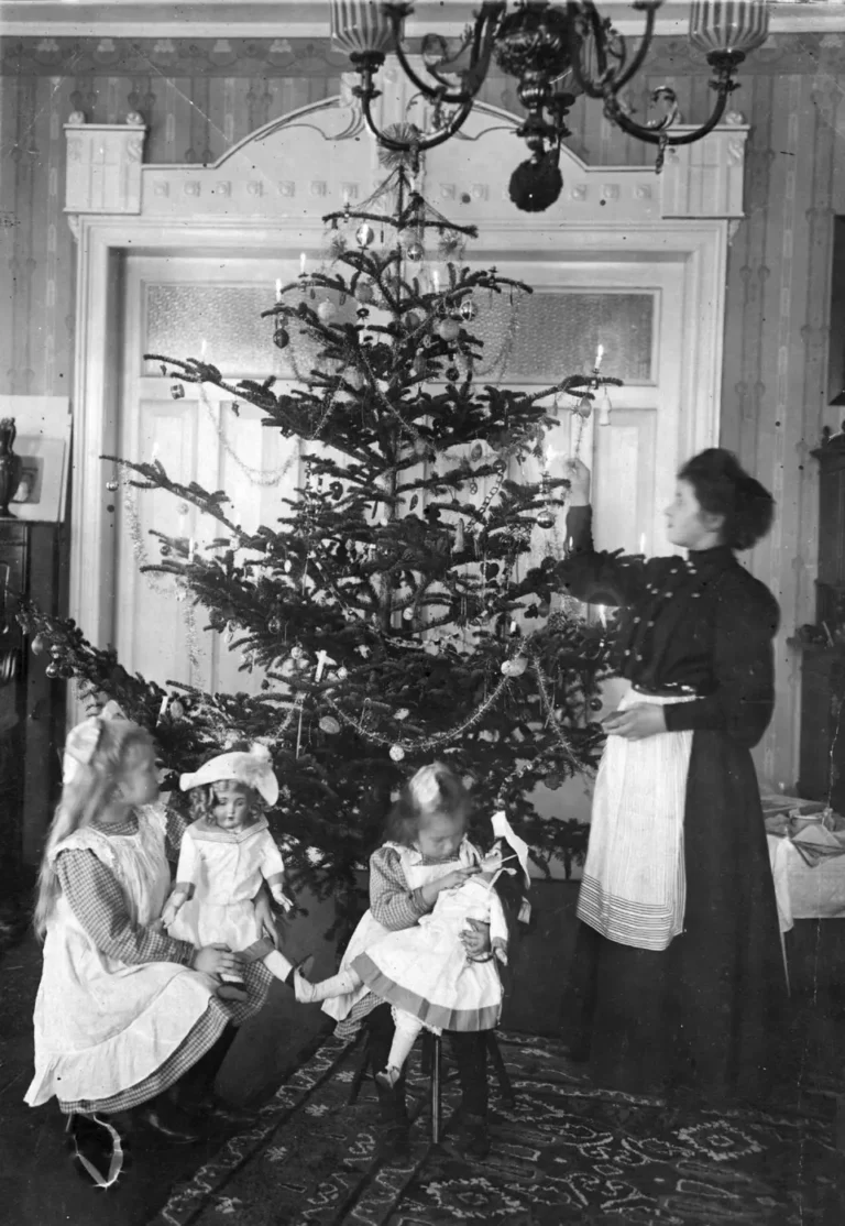 A black and white photo featuring two little girls in Edwardian dresses and their governess wearing a black Edwardian dress and a white apron lighting a candle on the Christmas tree.