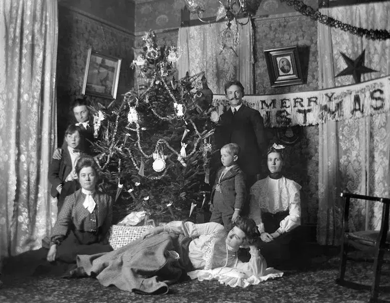 A black and white photo of a family in the late 19th century sitting around their Christmas tree, with a Merry Christmas sign, a star, a tinsel garland, ornate curtains, and framed photographs on the wall.