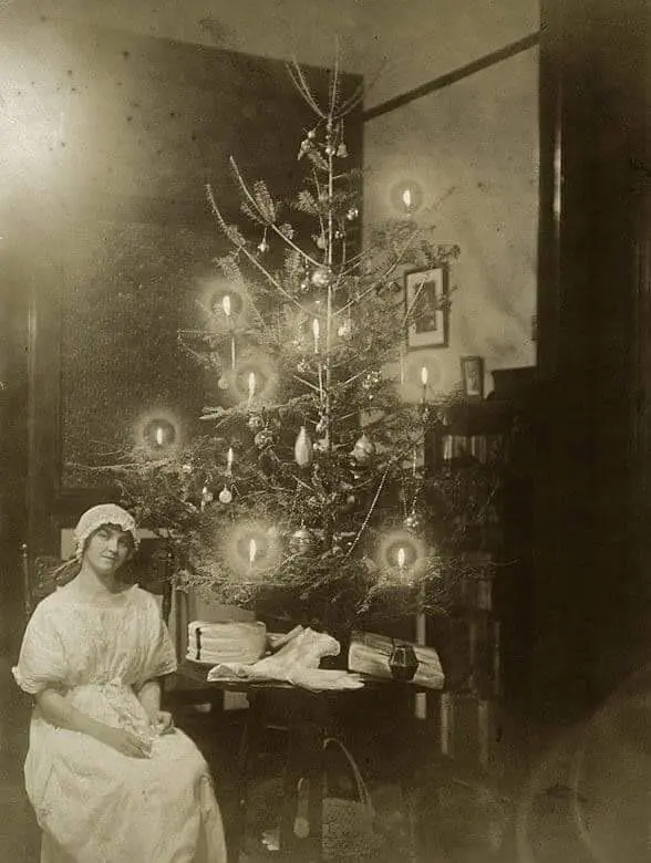 A black and white photograph of a woman dressed in white Edwardian clothing sitting next to a small Christmas tree placed on a table. The tree is decorated with candles and vintage ornaments.