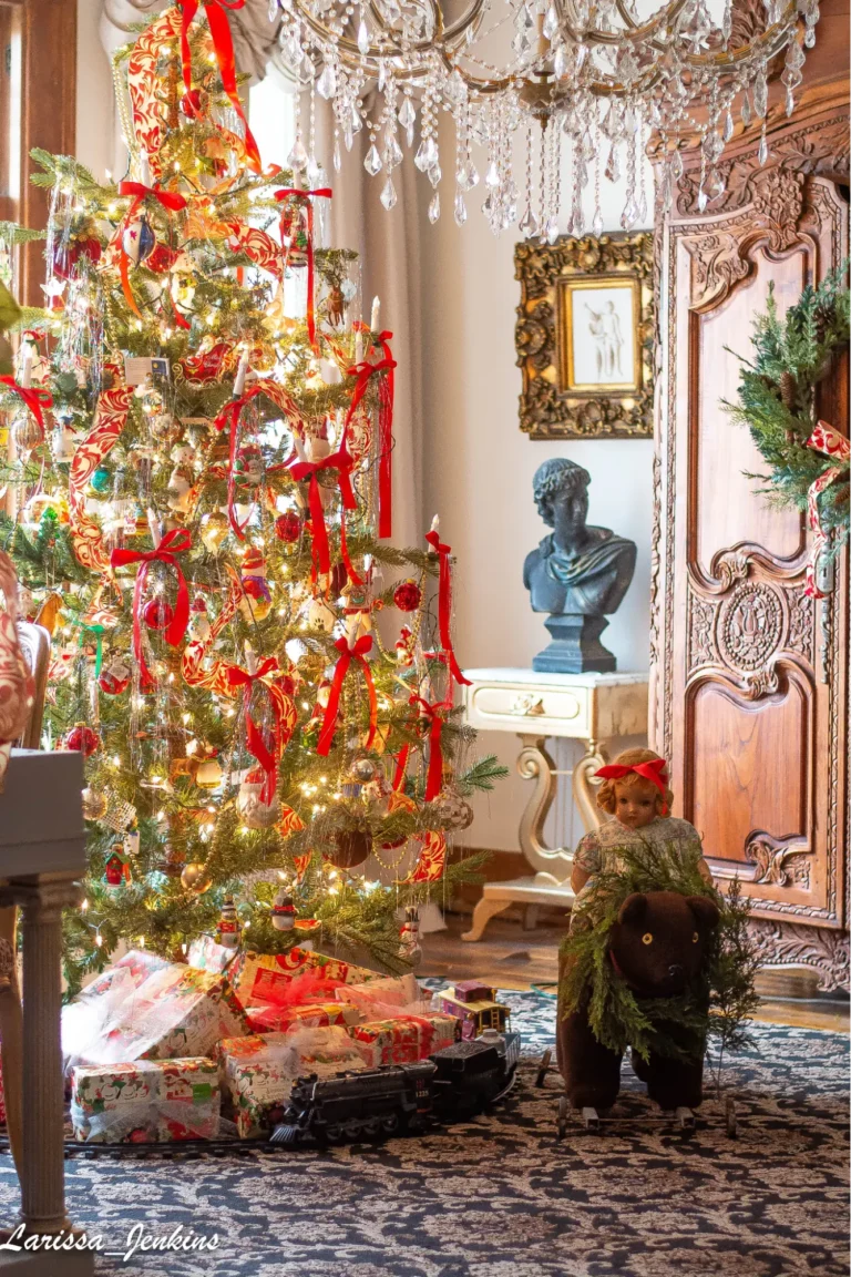 A large Christmas tree decorated with vintage ornaments, red bows, bead garlands, and string lights, with lots of presents underneath it, with an antique-style train set, and a vintage rocking bear with a doll sitting on it. The room also features a rug with an ornate pattern, an anthracite-colored bust, an antique ornate wooden wardrobe, a crystal chandelier, and an ornate gilded frame.