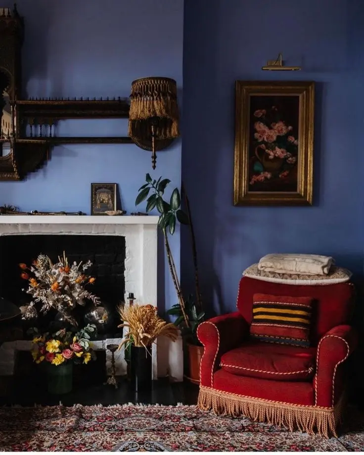 An antique interior with blue-colored walls, a vintage red armchair with a fringed bottom, a white brick fireplace mantle, dried flowers, and antique artwork