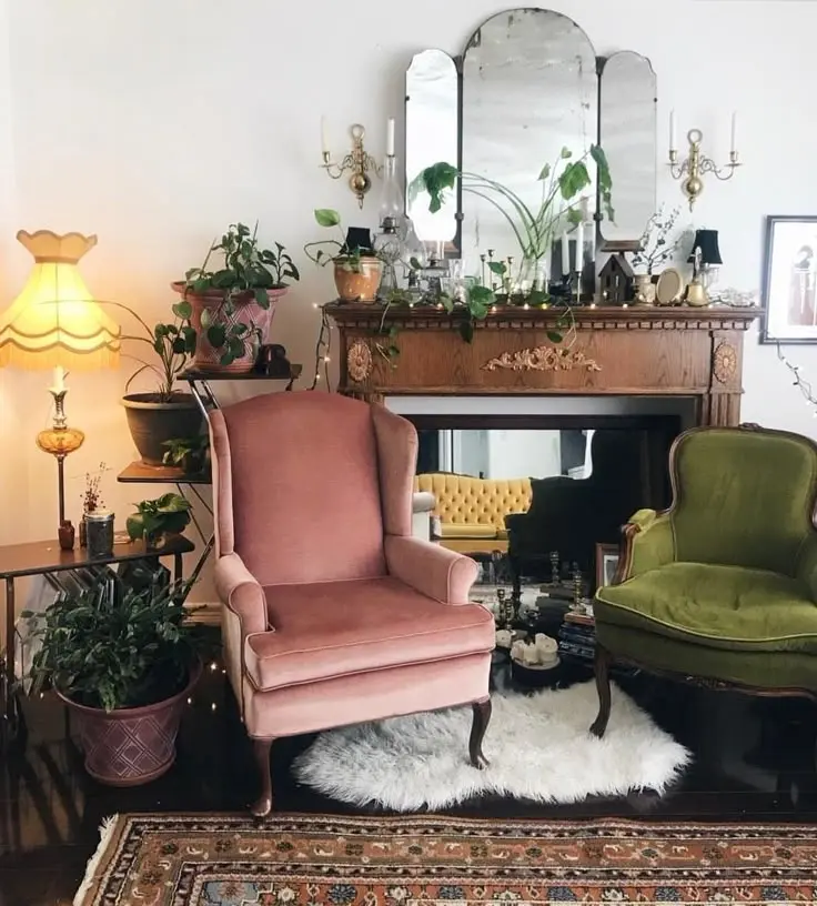 A vintage maximalist room with a vintage pink velvet armchair and a green vintage velvet armchair, fuzzy white area rug, vintage fringed standing lamp, plants, a mirror, and an antique fireplace mantel.