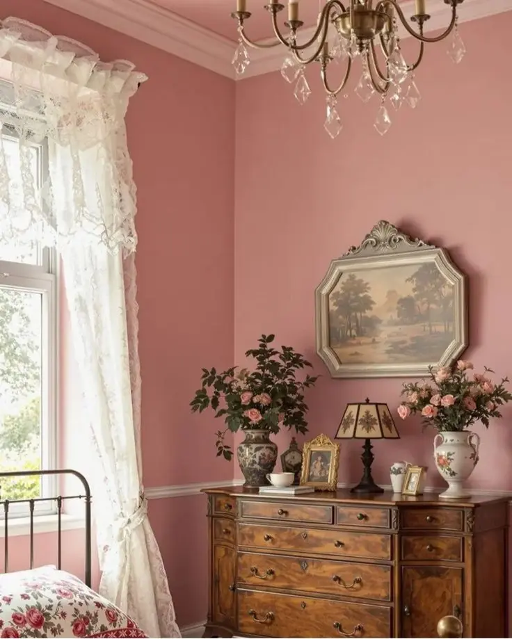 A cozy vintage bedroom with pink walls, an antique dresser, pink roses in vases, a window with lace curtains, and a landscape painting on the wall.